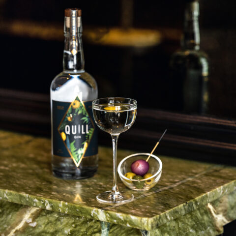 A small bowl of olives, a full pony glass and a bottle of gin on a green marble countertop.