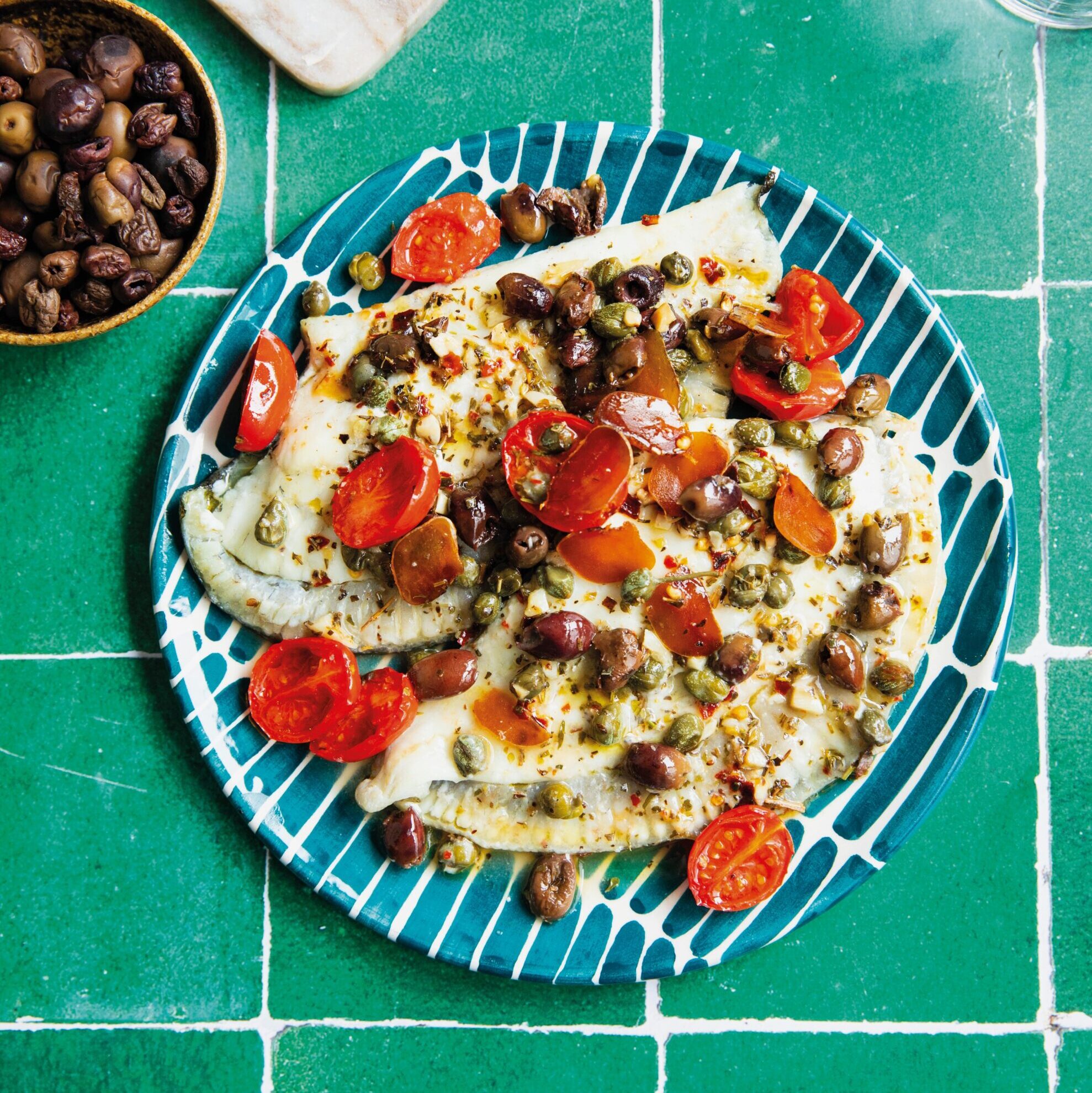 A striped plate with white fish topped with cherry tomatoes and capers.