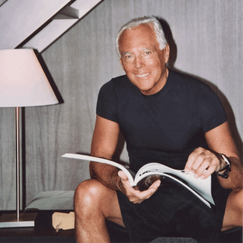 A tanned man with white hair smiling at the camera while holding a magazine.