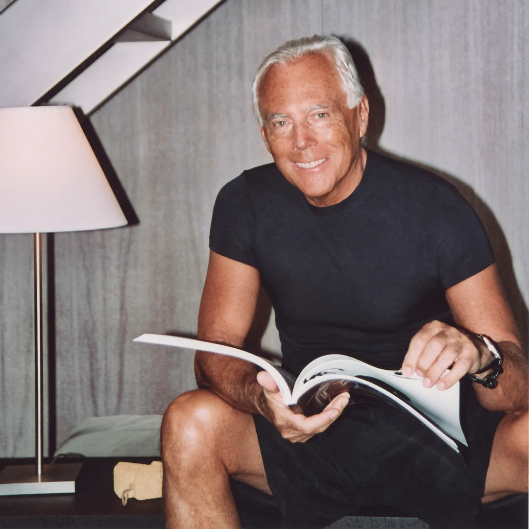 A tanned man with white hair smiling at the camera while holding a magazine.