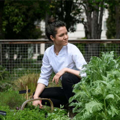 A woman in a white chef's coat crouching in a garden.