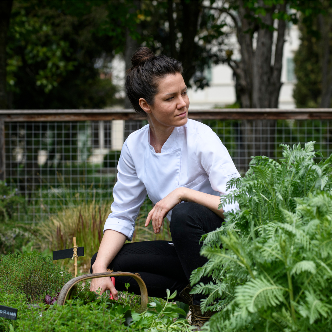 A woman in a white chef's coat crouching in a garden.