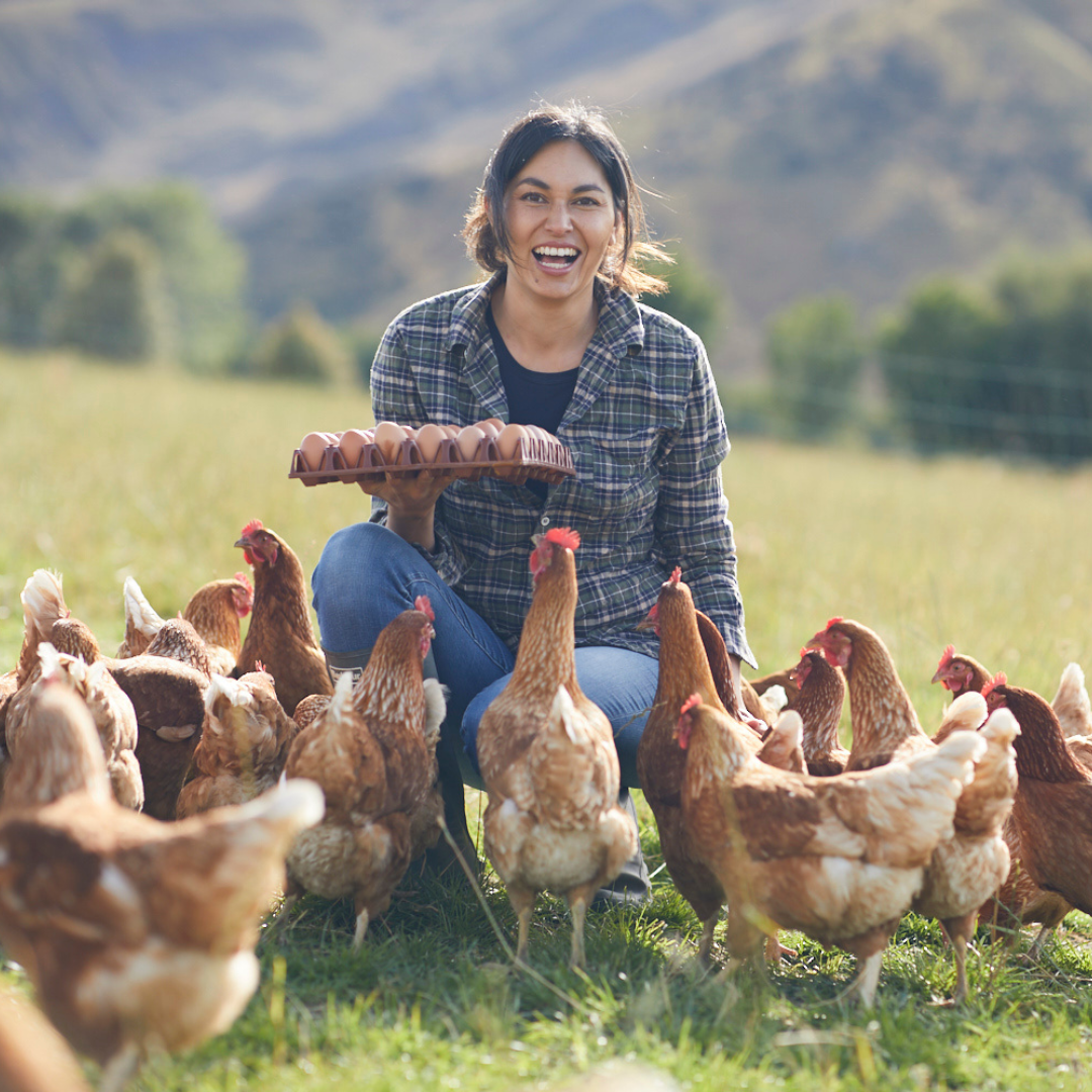 A smiling woman holding a carton of eggs in an open field, surrounded by chickens.