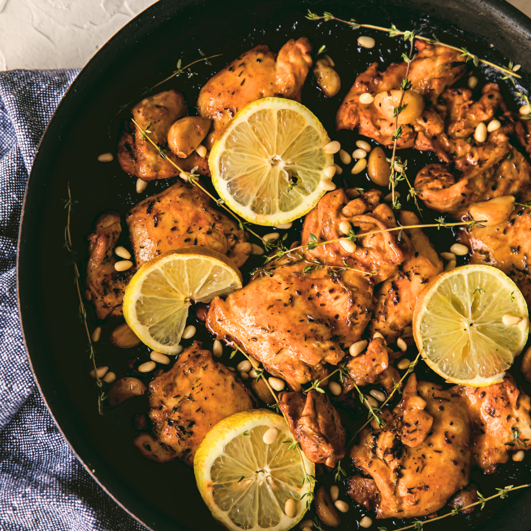 A close-up of a black pan with cooked chicken thighs in a brown sauce, topped with pine nuts and lemon slices.