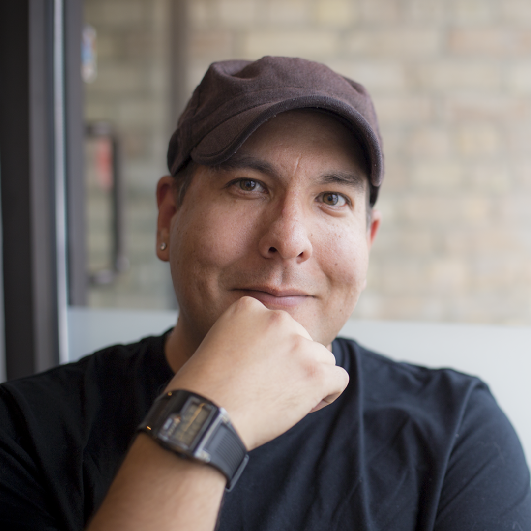 A headshot of a smiling man wearing a hat, resting his hand on his chin.