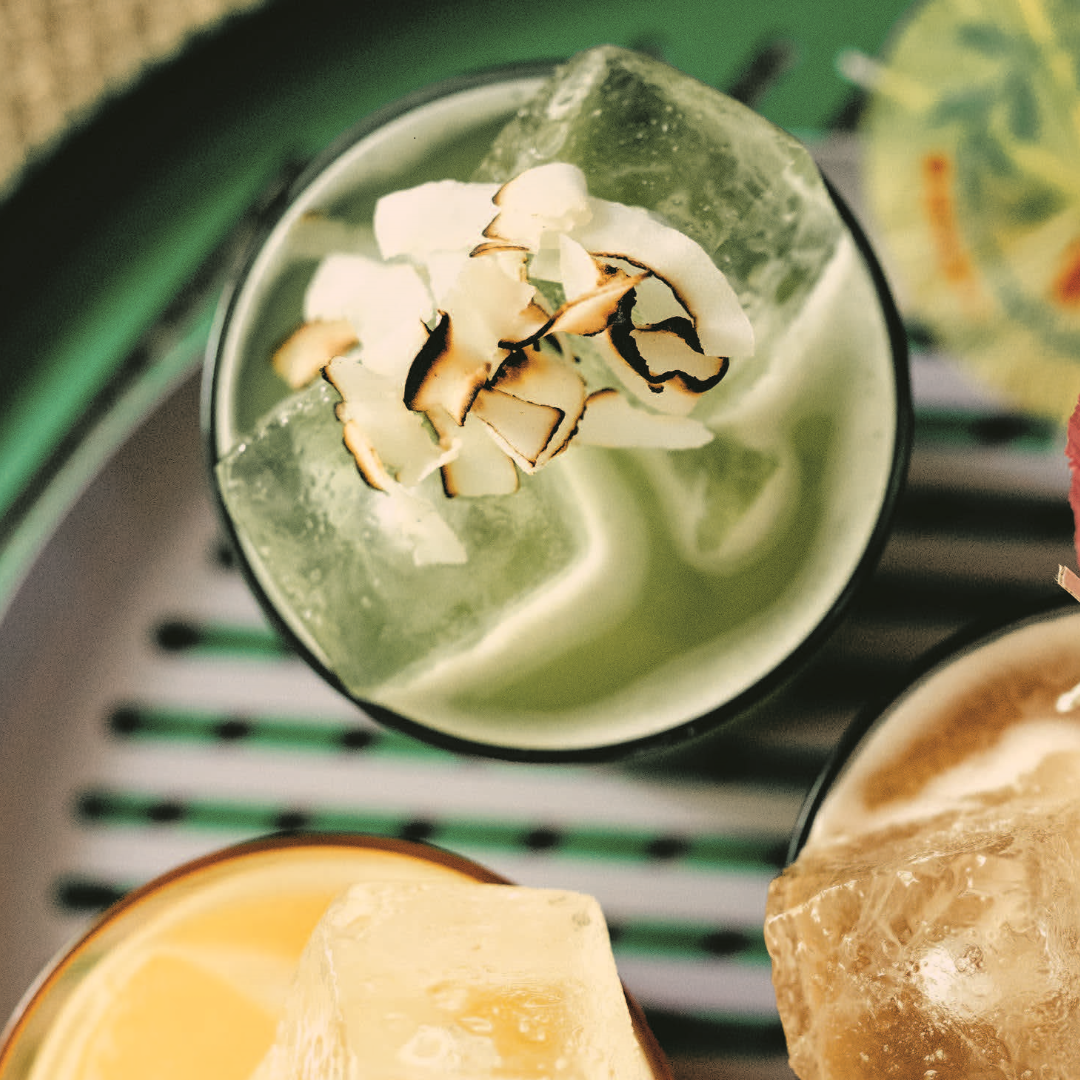 A top angle close-up shot of a glass with a green liquid, ice cubes and toasted coconut flakes.