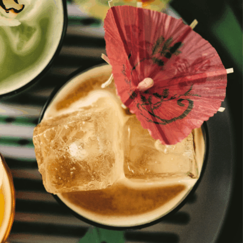 A top angle close-up shot of a glass with two pieces of ice, yellow liquid and a decorative pink drink umbrella.