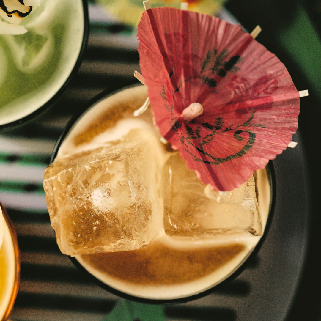 A top angle close-up shot of a glass with two pieces of ice, yellow liquid and a decorative pink drink umbrella.