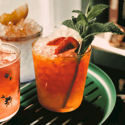 A glass filled with pebble ice, red liquid, and garnished with strawberry slices and a sprig of mint.