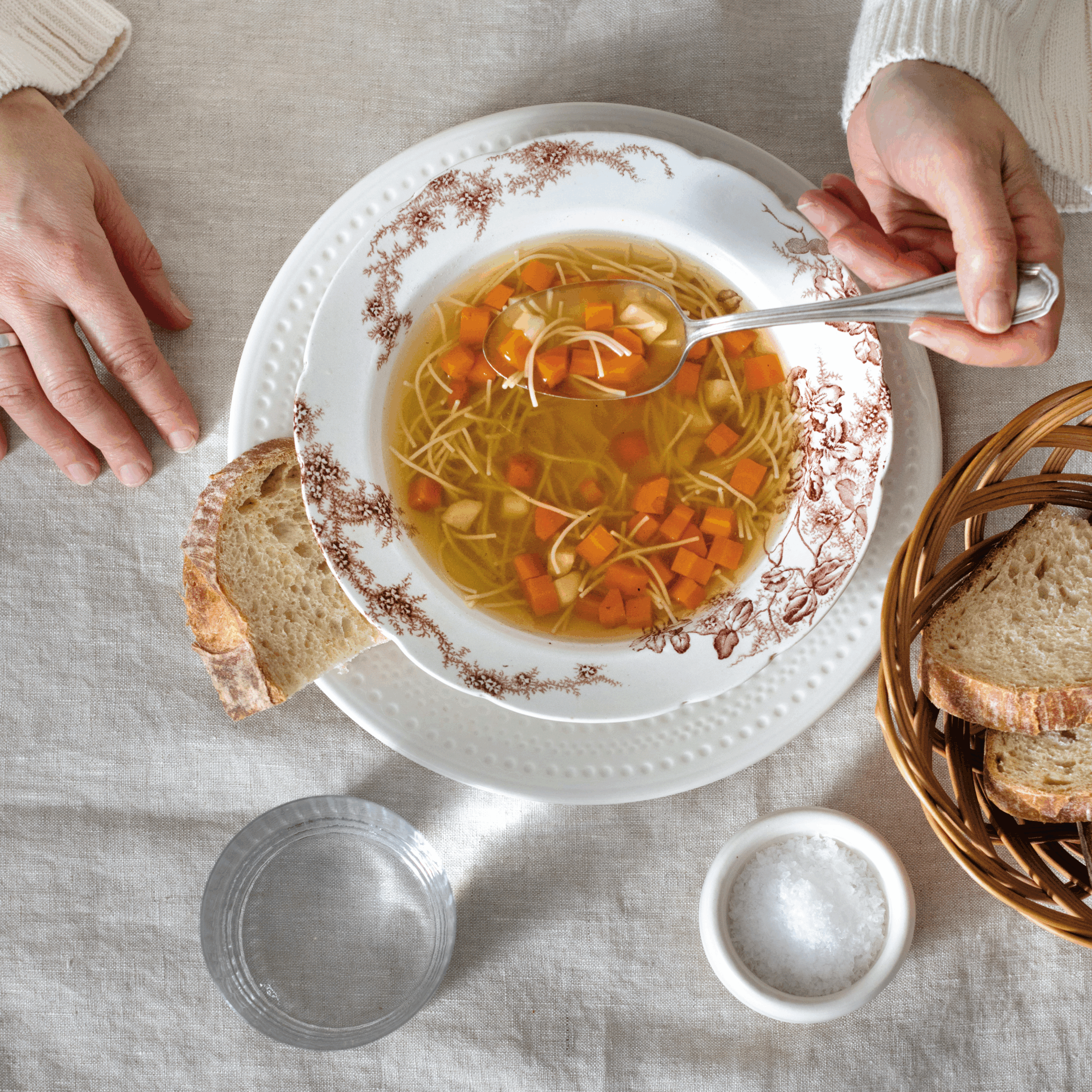 One hand holding a spoon in a bowl of chicken noodle soup, with a piece of bread next to it.