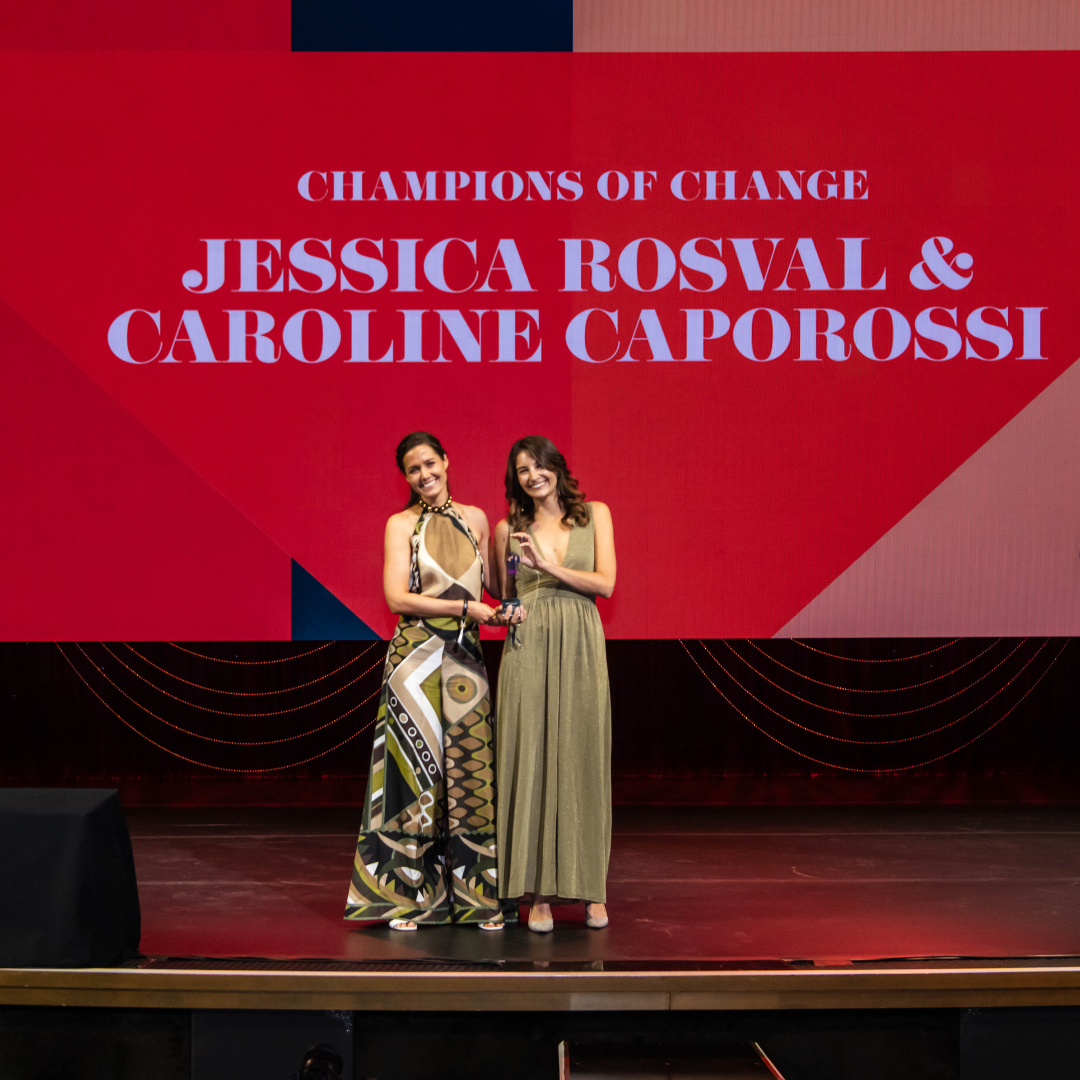 Two women on stage accepting an award with a red screen that lists their names.