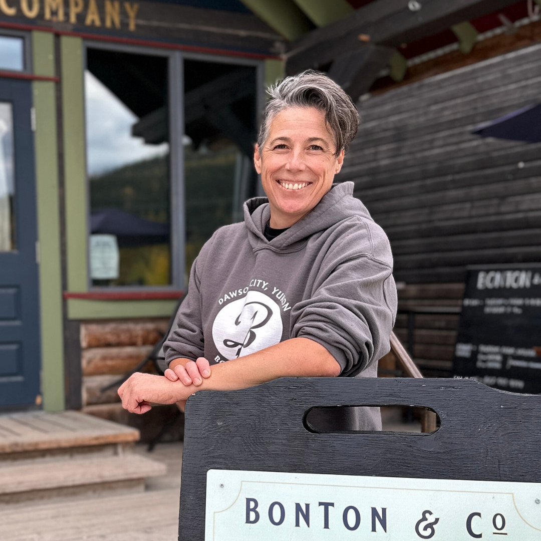 A woman with short gray hair in a gray hoodie in front of an outdoor sign.