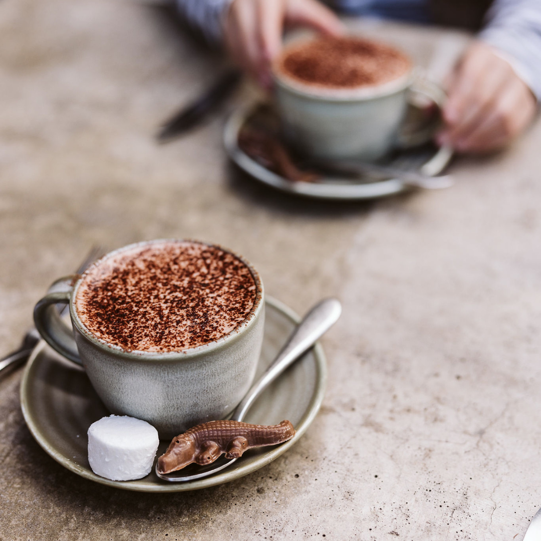A cup of hot chocolate with a spoon, a marshmallow and a crocodile-shaped chocolate on the saucer.