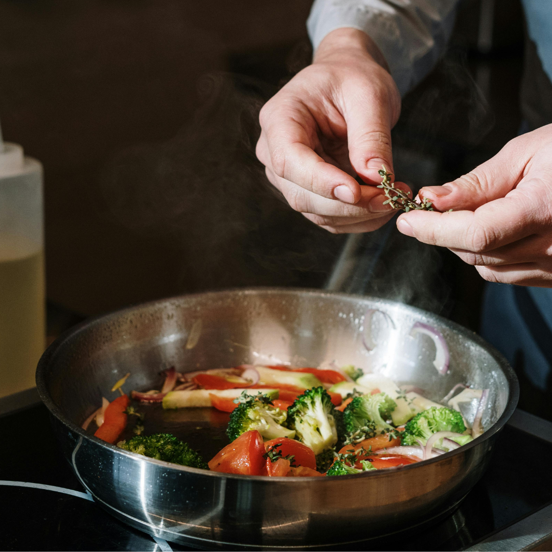 Hands holding an herb over a pan filled with broccoli and red peppers on an induction stovetop.