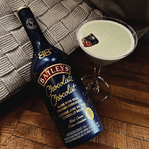 A martini glass filled with a foamy light green drink next to a dark blue bottle.