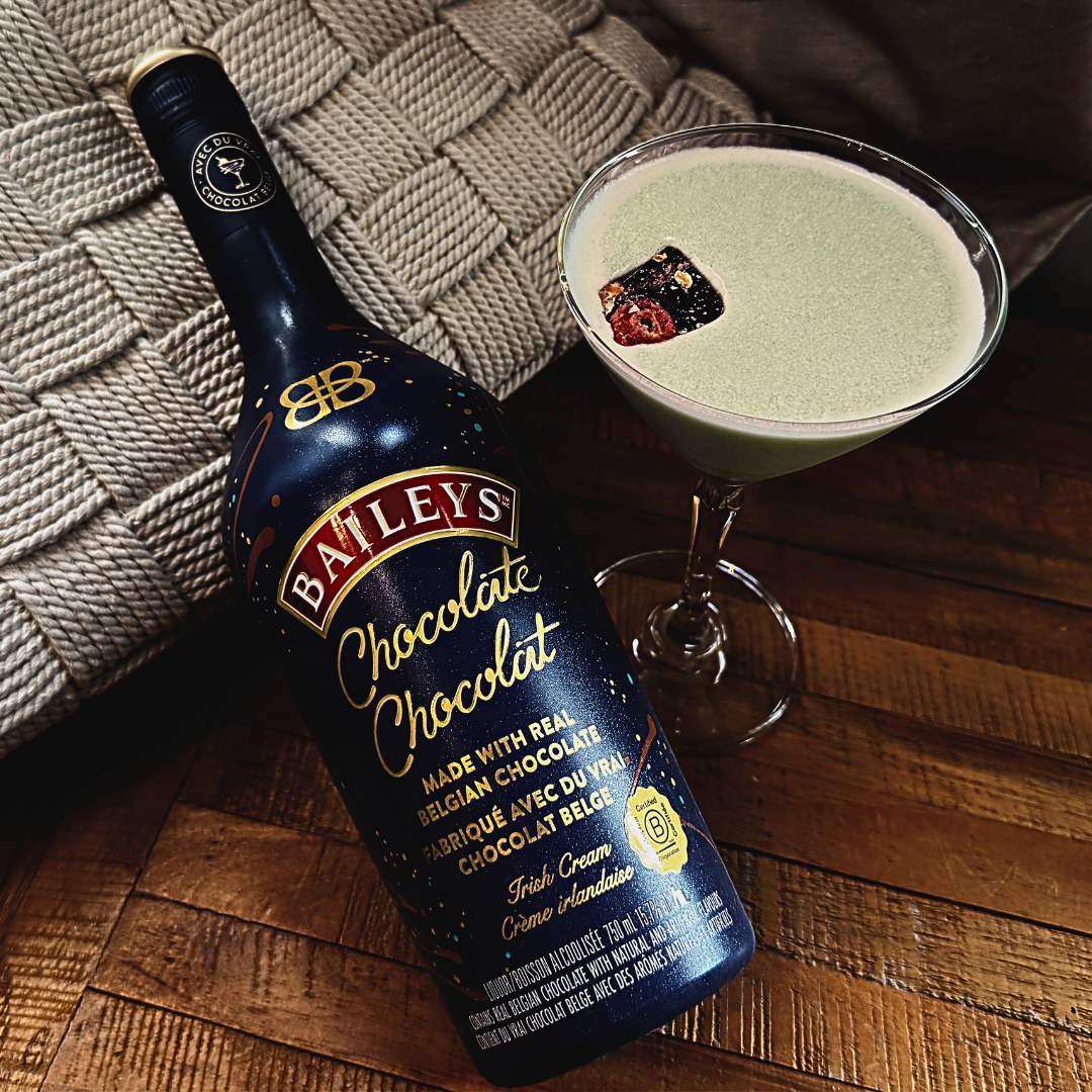 A martini glass filled with a foamy light green drink next to a dark blue bottle.