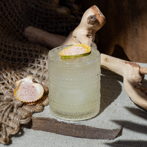 A margarita topped with a lime wheel on a sandy surface with a seashell.