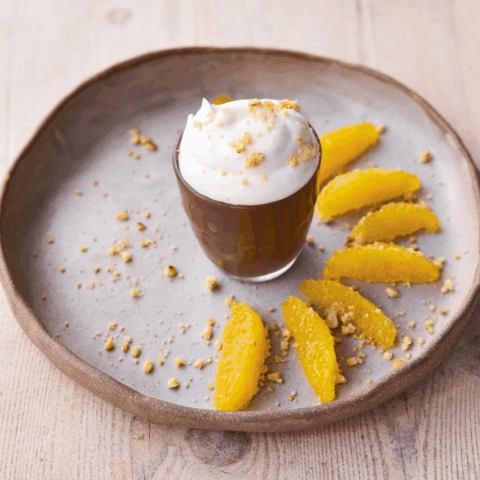A small chocolate pot filled with Greek yogurt and chopped hazelnuts with six slices of orange on a plate.