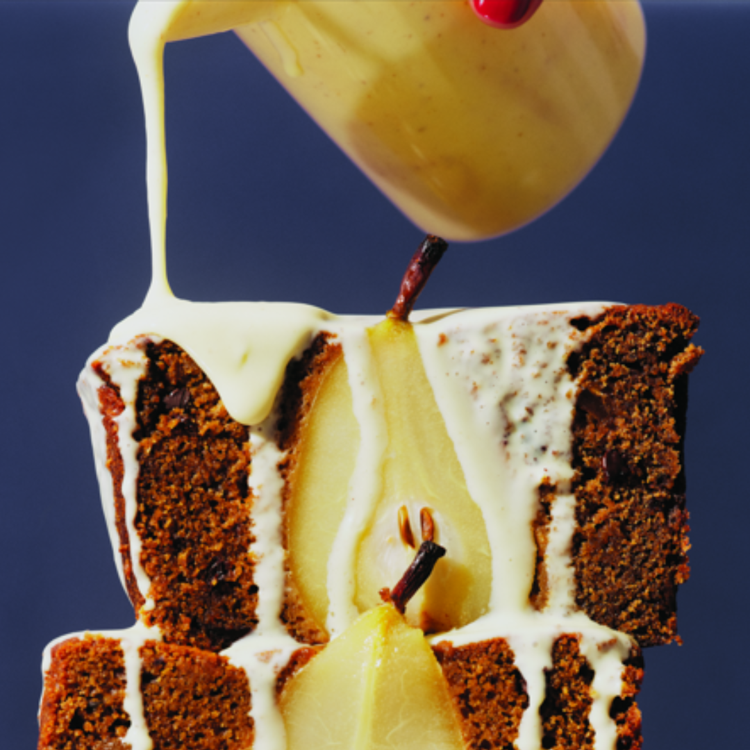 Thick cream being poured onto two slices of brown loaf cake with a pear in the centre.