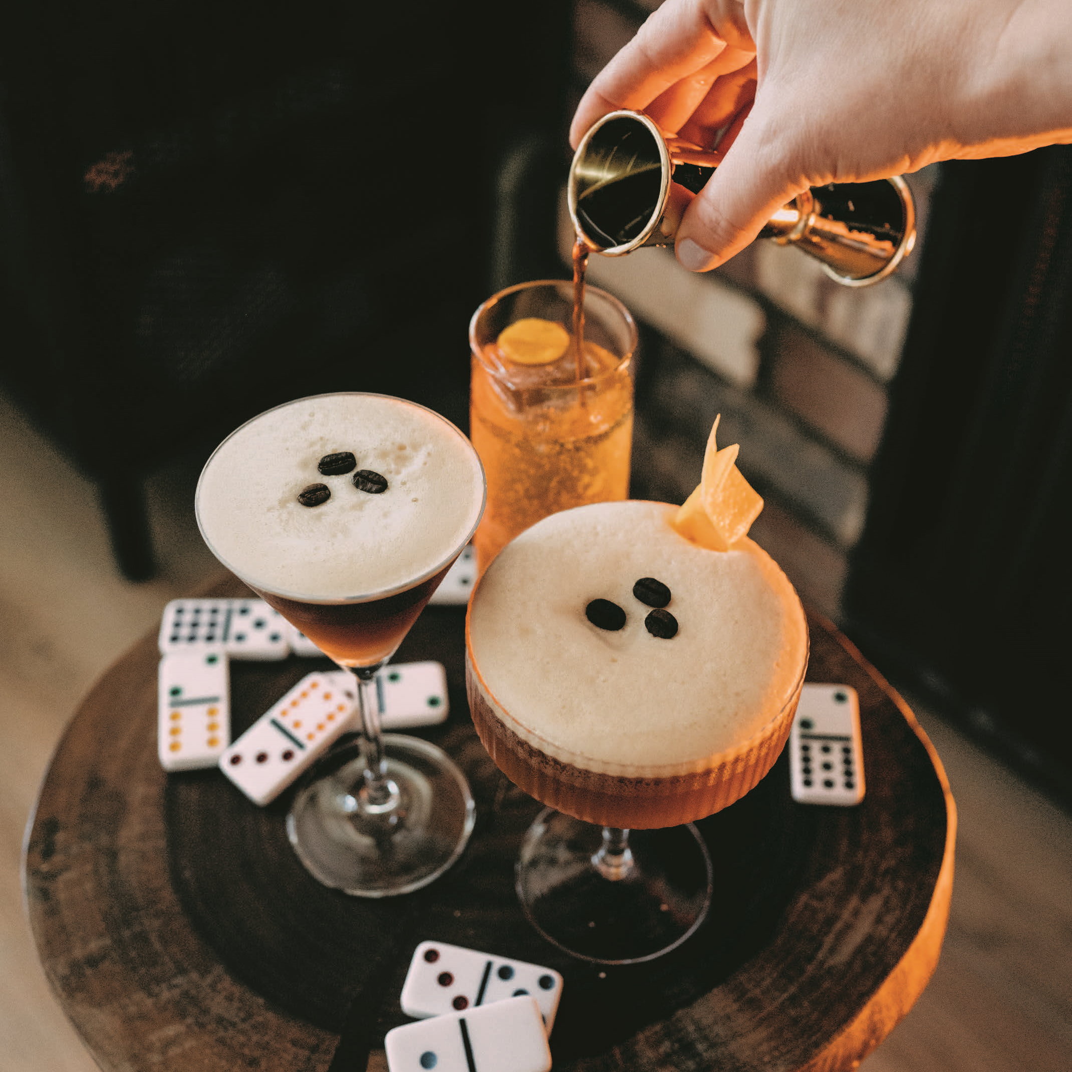 Hand pouring liquid from a jigger into a glass on a tray with two espresso martinis.