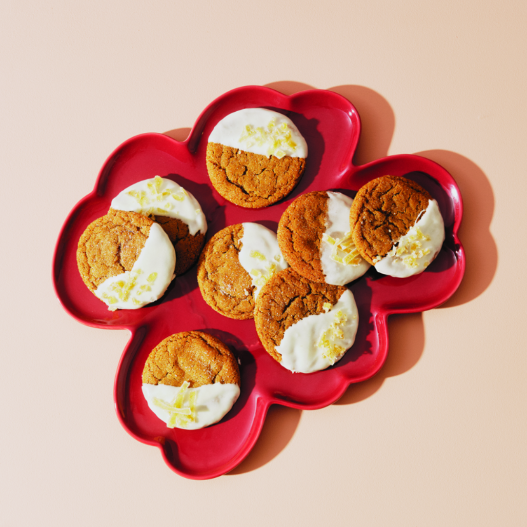 Eight ginger cookies half-dipped in white frosting and garnished with candied ginger on a red plate.