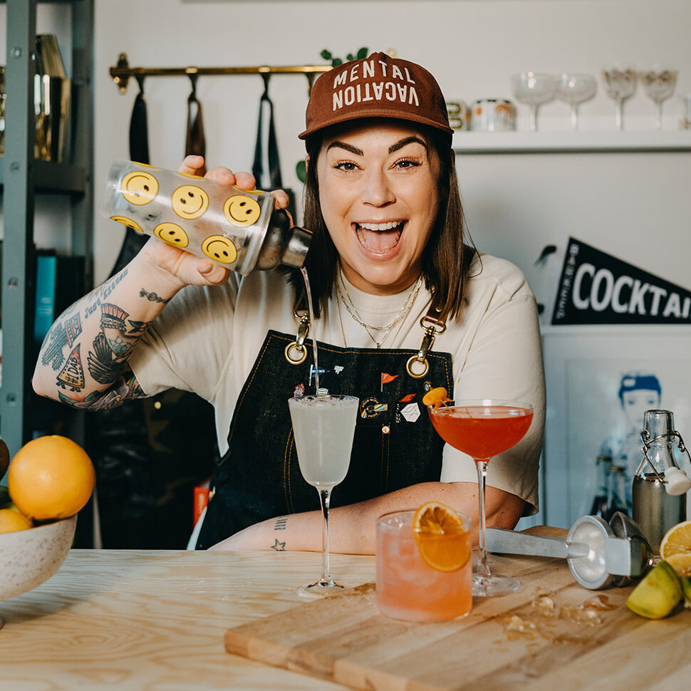Smiling woman in brown baseball cap pours clear liquid from a shaker into a glass.