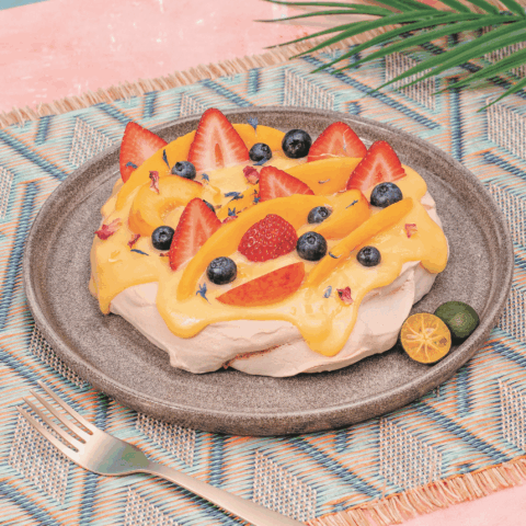 Pavlova topped with an orange sauce, strawberries, blueberries, peach slices next to sliced calamansi on a plate.