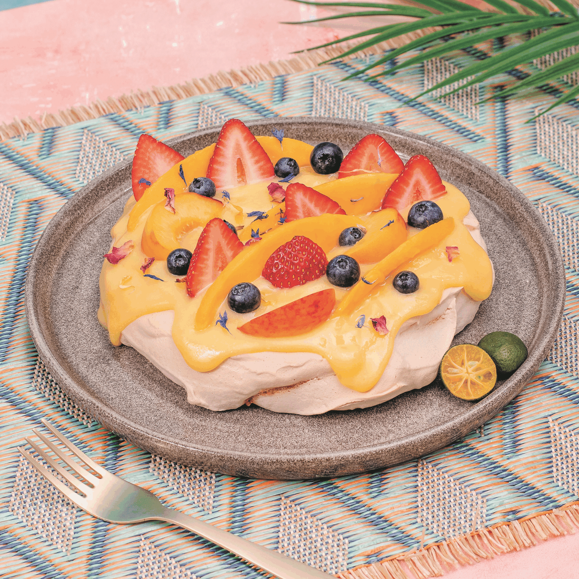 Pavlova topped with an orange sauce, strawberries, blueberries, peach slices next to sliced calamansi on a plate.