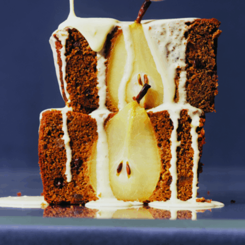 Two slices of brown loaf cake with half a pear inside being drizzled with runny white sauce.
