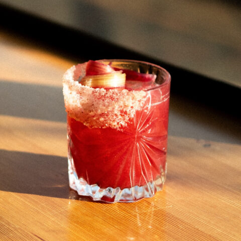 A red rhubarb margarita in a salt-rimmed glass garnished with a rhubarb swirl.