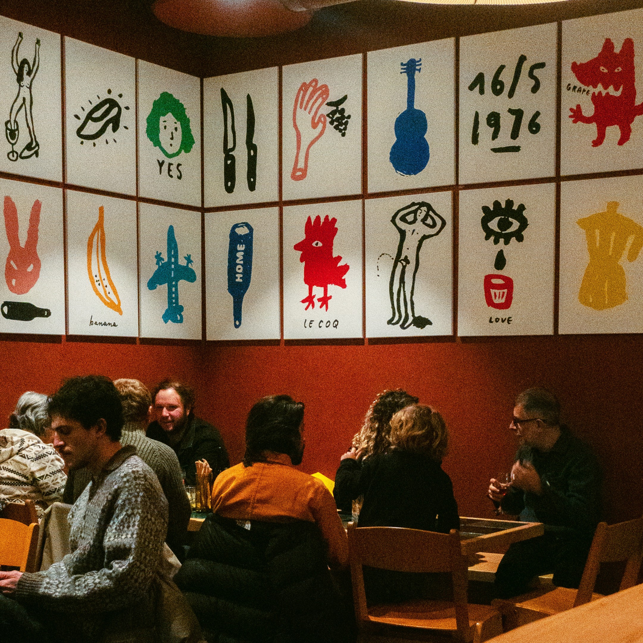 Red-toned dining room with wooden tables and illustrated panels on the walls, people seated enjoying meals and drinks.