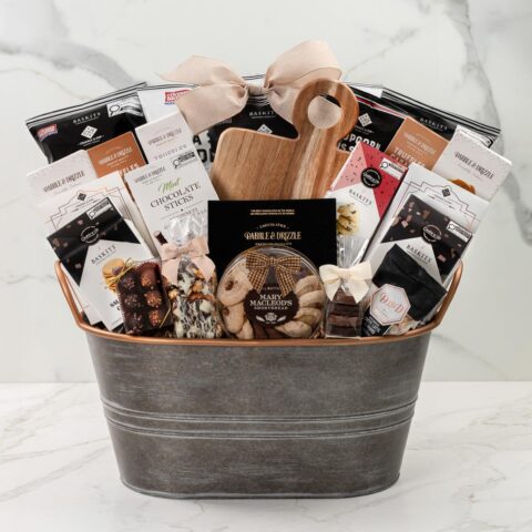 A gift basket featuring chocolates, treats and a wooden cutting board in a metal bucket on a marble background.