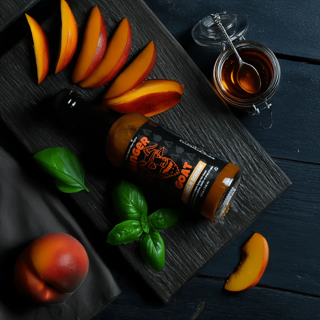 A bottle of hot sauce next to a jar of maple syrup and peaches on a black wooden board.