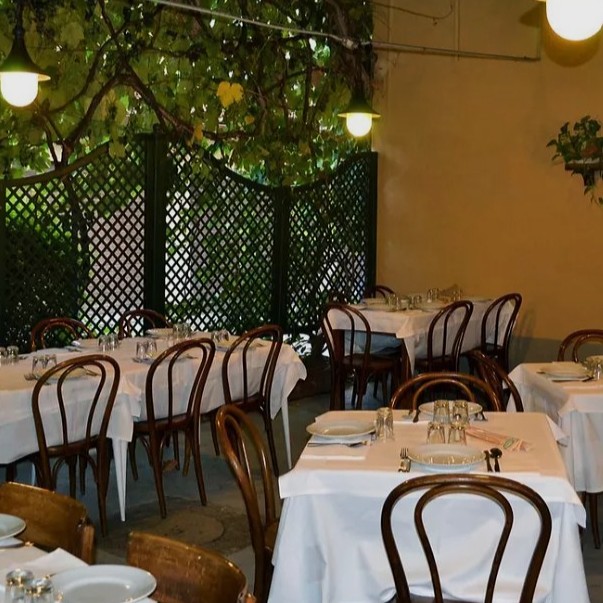White tables and wooden chairs set up with lights overhead and greenery.