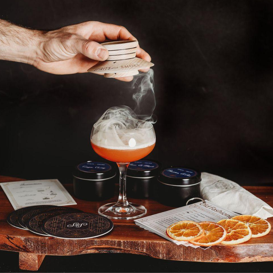 A hand holding a smoking saucer above a cocktail glass with rising smoke.