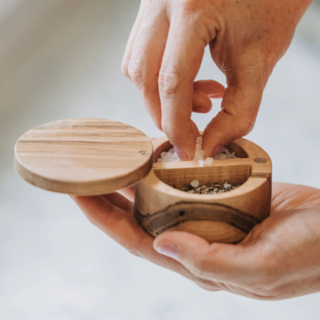 A hand grabbing salt from a wooden salt cellar with a magnetic lid and two compartments.