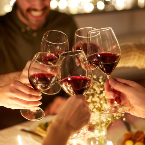 Man smiling in the background as five glasses of red wine come together to cheers at this Tuscany 12 days of wine pairings