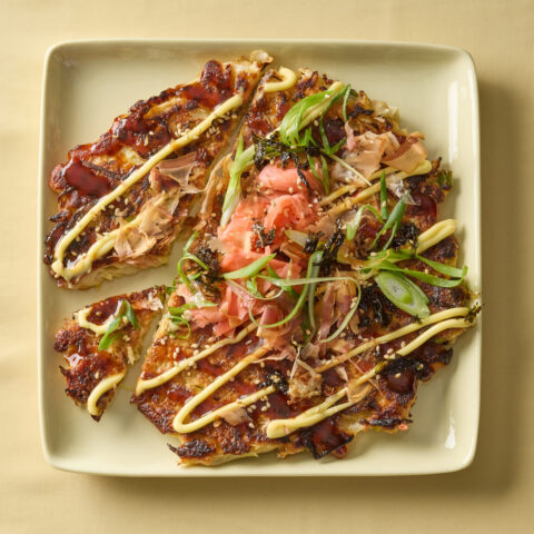 Okonomiyaki, a crisp golden, Japanese pancake, cut into wedges on a square plate, topped with mayonnaise, sweet sauce, scallions, bonito flakes, seaweed and pickled ginger.