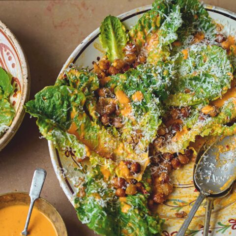 Romaine lettuce tossed with creamy orange dressing and crispy spiced chickpeas, topped with grated cheese, served on a plate with spoons and extra dressing on the side.