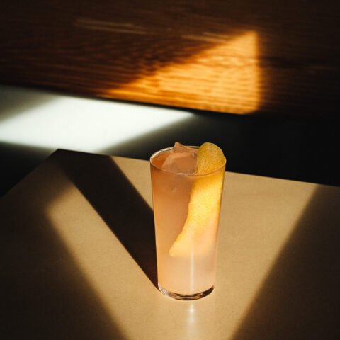 Tall glass of a pale pink cocktail with ice and a citrus peel, set on a table in dramatic sunlight with sharp shadows casting a geometric pattern around it.