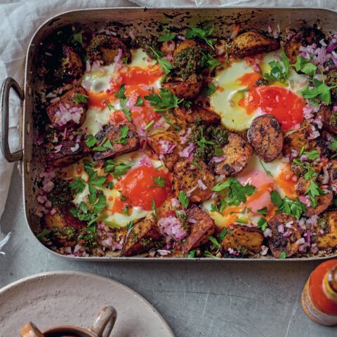 Roasted potatoes baked with eggs in a pan, topped with herbs, chopped red onion and green chermoula sauce, served with coffee and utensils on a table.