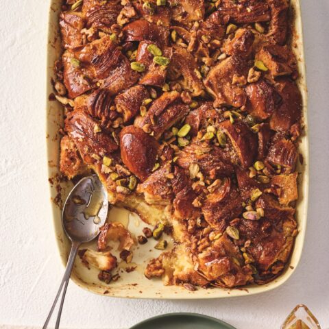 Baked French toast topped with chopped nuts and syrup in a dish, with a serving on a plate and a small pitcher of syrup alongside.