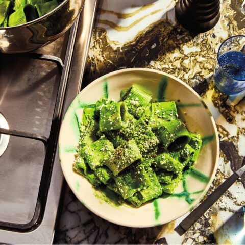 Tube-shaped pasta coated in a bright green sauce, topped with grated cheese and black pepper, served in a bowl beside a pot on a stovetop.