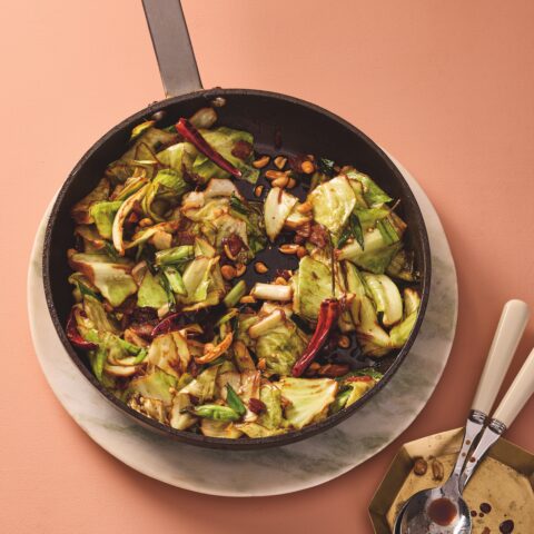 Cast-iron skillet filled with hot and sour cabbage stir-fry, tossed with dried chilies, peanuts and scallions, set on a soft peach background with serving spoons nearby.