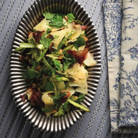 Shaved asparagus salad with prosciutto, shaved cheese, fresh herbs and toasted nuts, served on a dark oval platter beside a patterned cloth napkin.