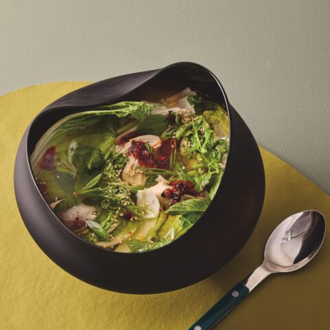 Modern black bowl filled with clear chicken and cabbage soup, topped with fresh herbs, chili oil and sesame seeds, set beside a spoon on a yellow placemat.
