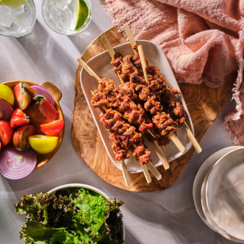 A plate of veal kabobs next to a bowl of lettuce and plate of onions and tomatoes.